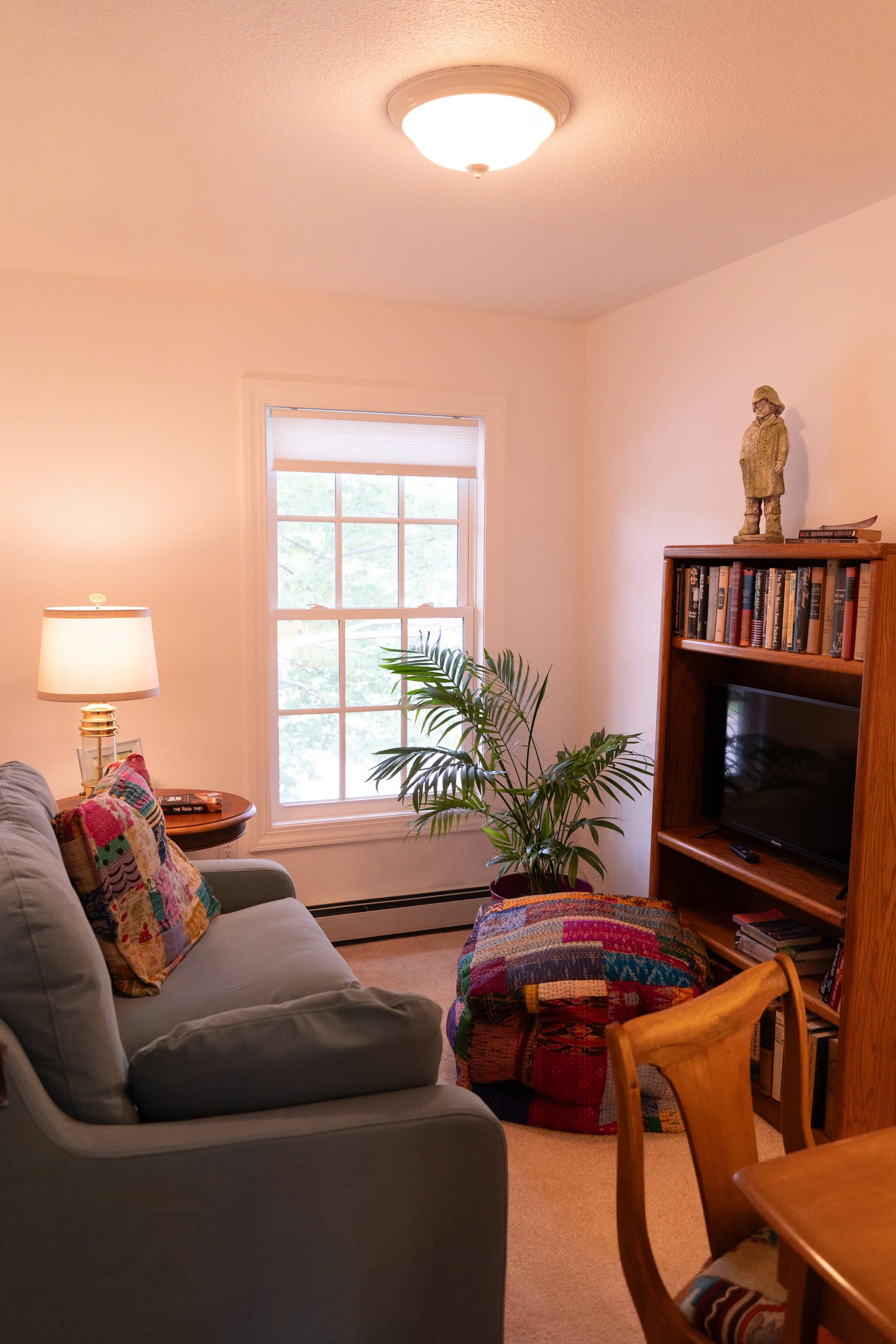Living room with a beige sofa, colorful throw pillows, a lamp, a window with blinds, a potted plant, a wooden bookshelf with books and a statue on top, a stained quilted ottoman, and a wooden dining chair.