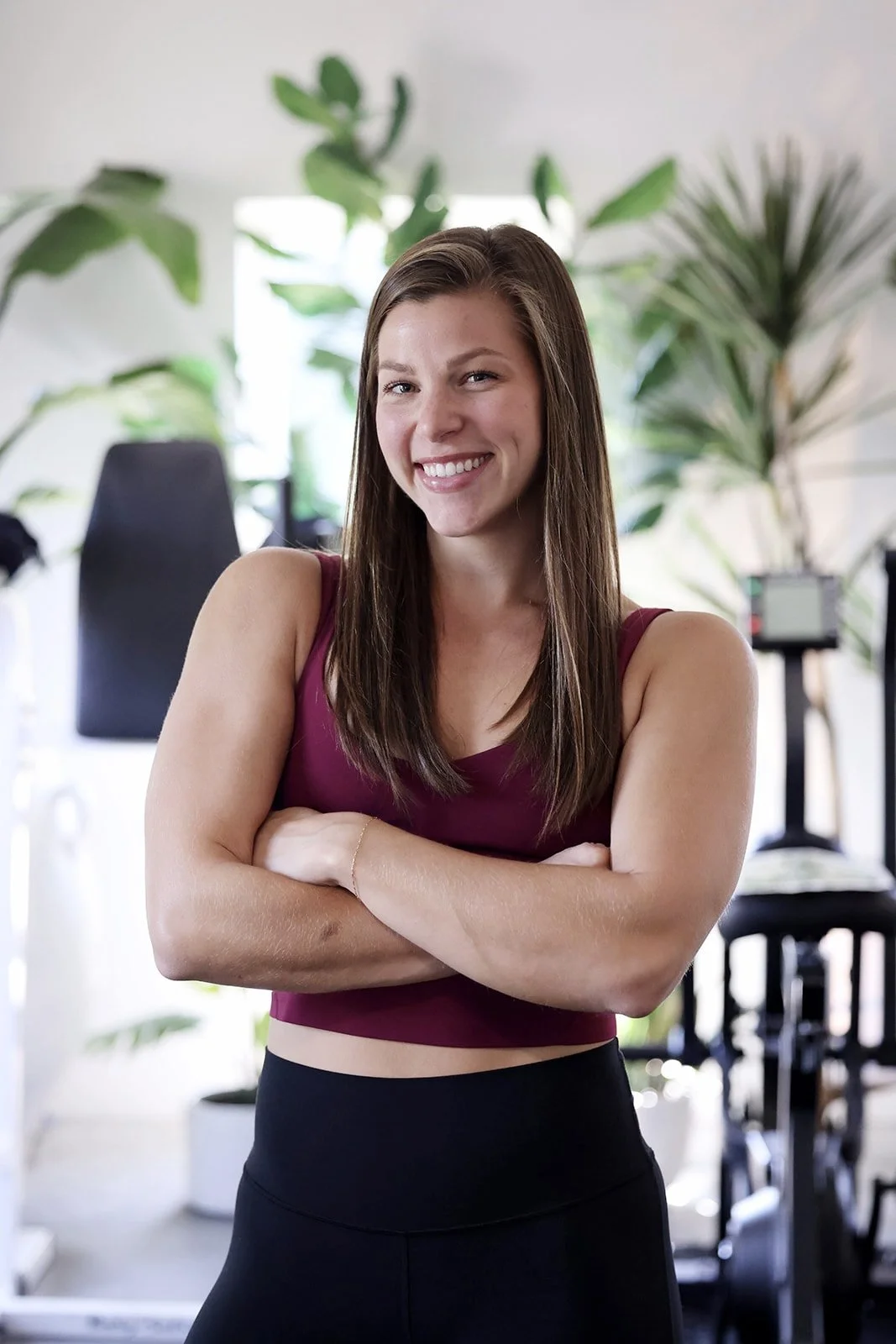 A smiling woman with long brown hair, wearing a sleeveless maroon top and black leggings, standing with arms crossed in a room with indoor plants and workout equipment.