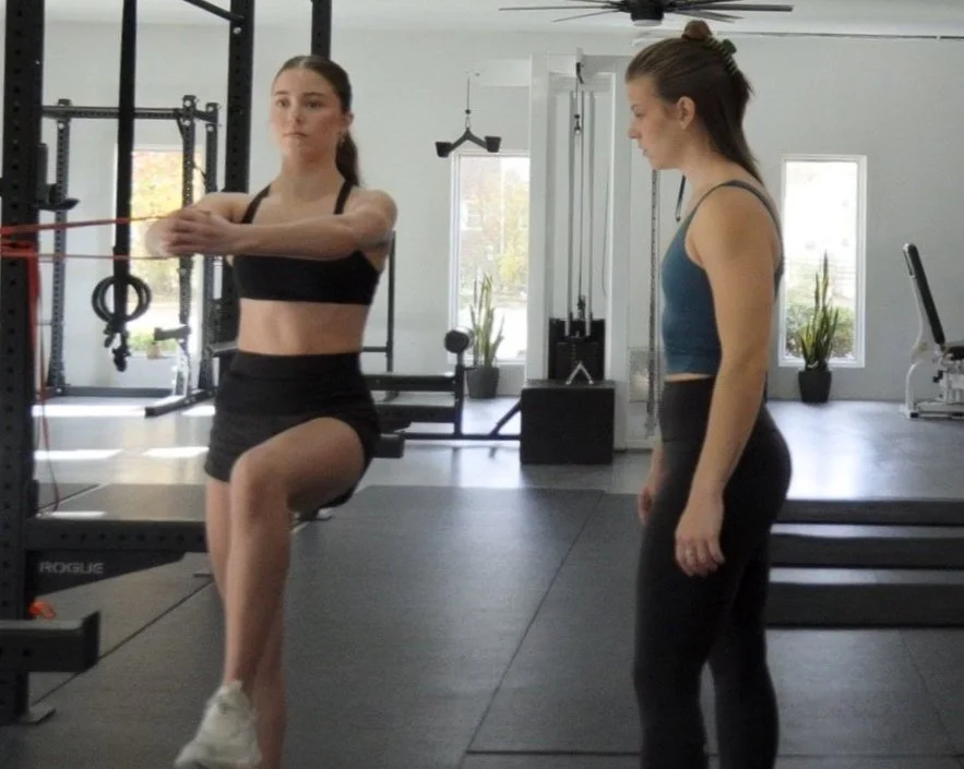 Woman performing a resistance band exercise with a trainer observing in a gym.