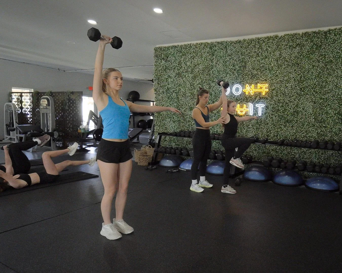 Four young women working out in a gym, using dumbbells and balance exercises, with a green leafy wall and a neon sign in the background.