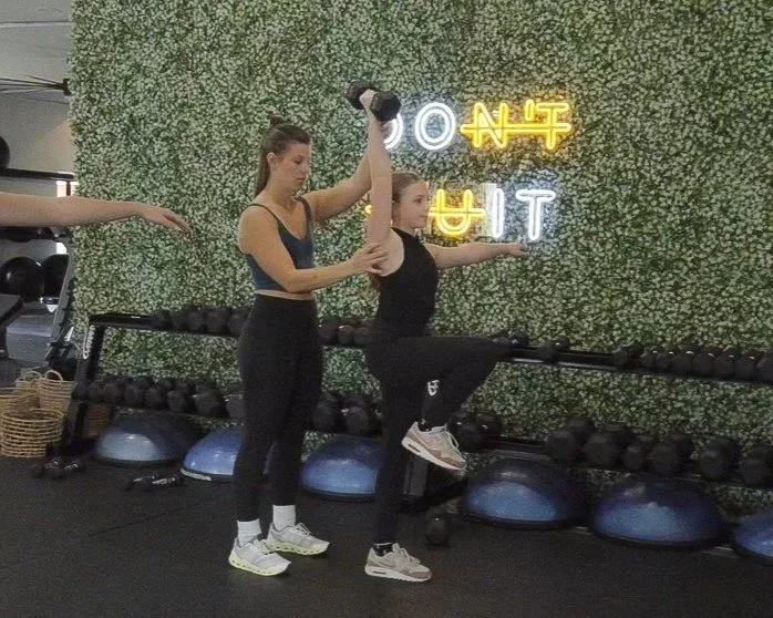 A woman in gym attire assisting a young girl with a workout in a gym with a green leafy wall and neon signs in the background.
