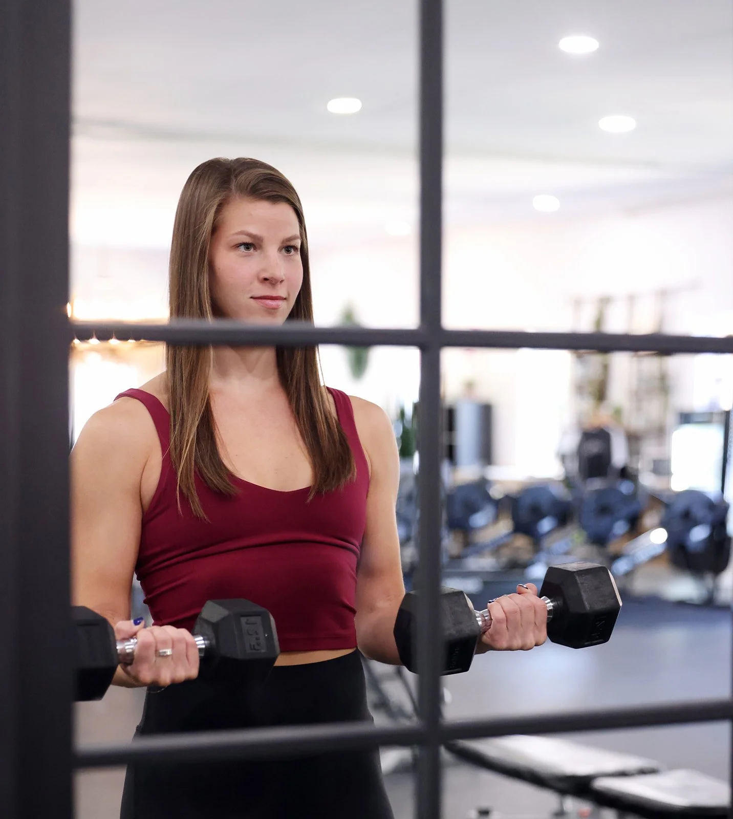 A young woman with long brown hair, wearing a maroon sports top and black workout pants, is lifting dumbbells in a gym, viewed through a window divided into panes.