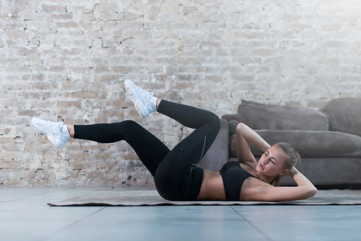 Woman exercising on a yoga mat in a living room with brick wall background, lying on her back with knees bent and hands behind head.