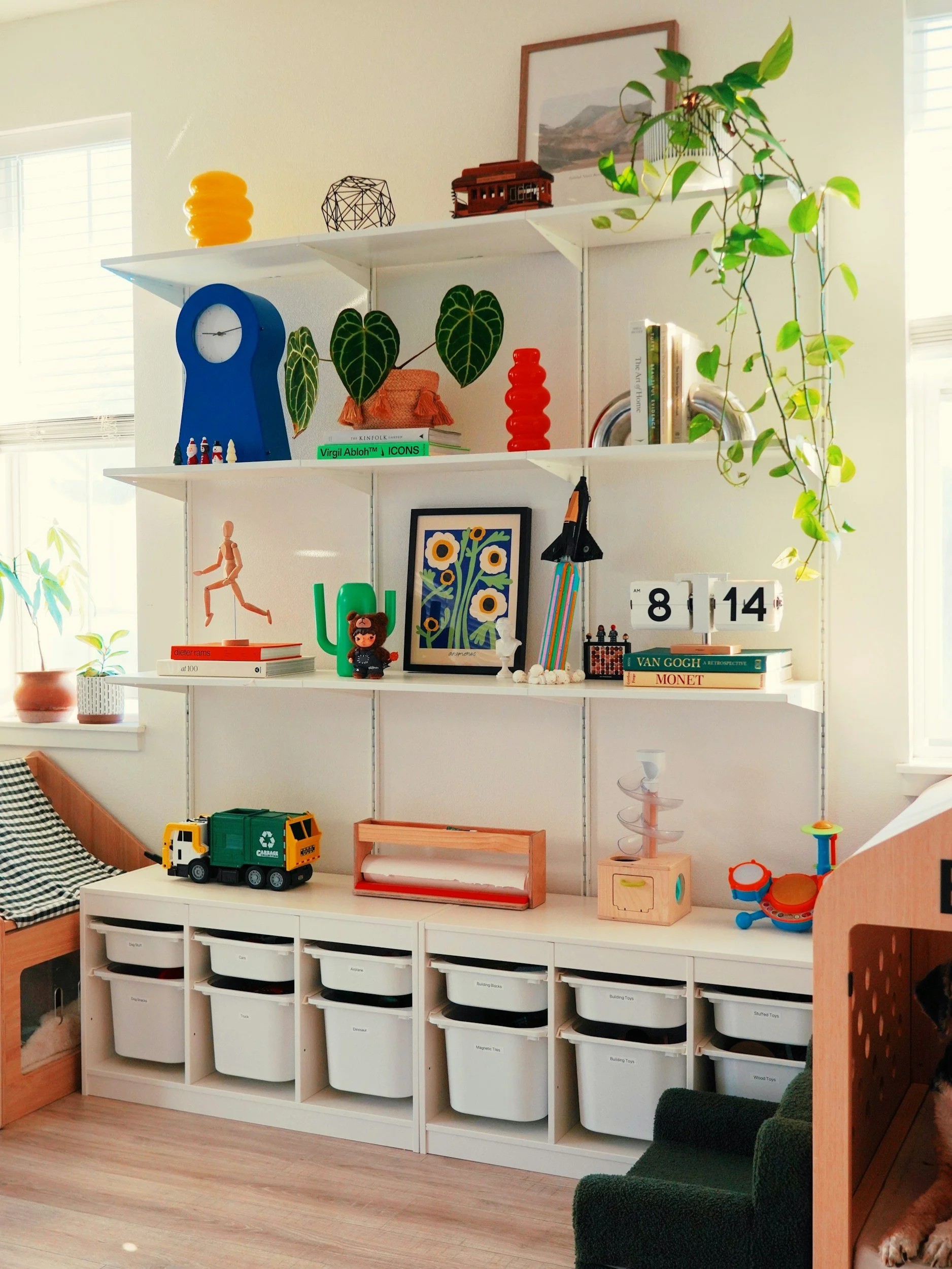 A white shelving unit with decorative items, books, and toys in a well-lit room. The shelves feature plants, art, colorful vases, a model train, and a clock calendar. The bottom section has labeled white storage bins. A window with blinds is on the left, and part of a green chair and a dog are visible.