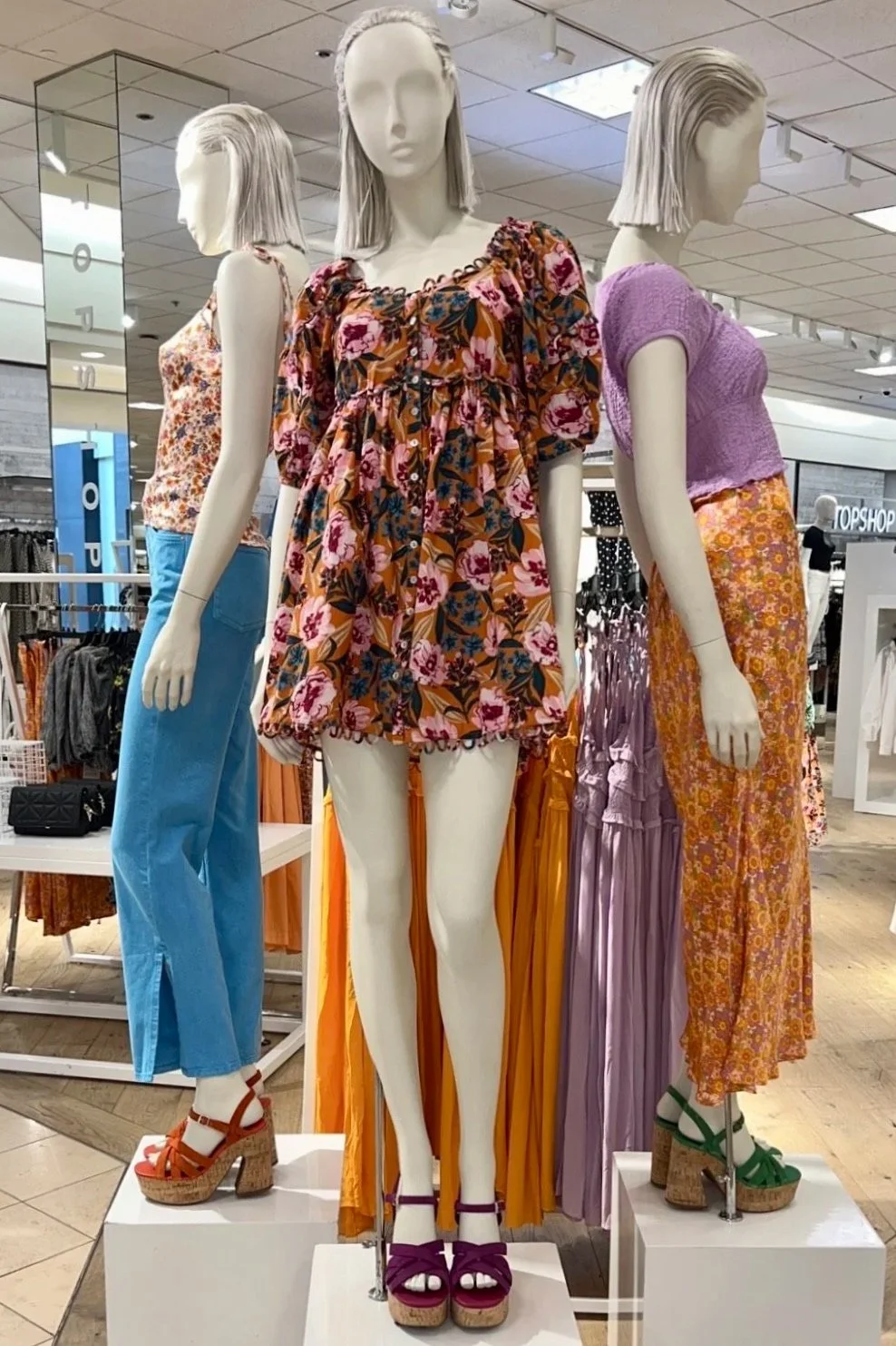 Three mannequins dressed in colorful, summery outfits displayed in a retail store, with clothing racks and store signage visible in the background.