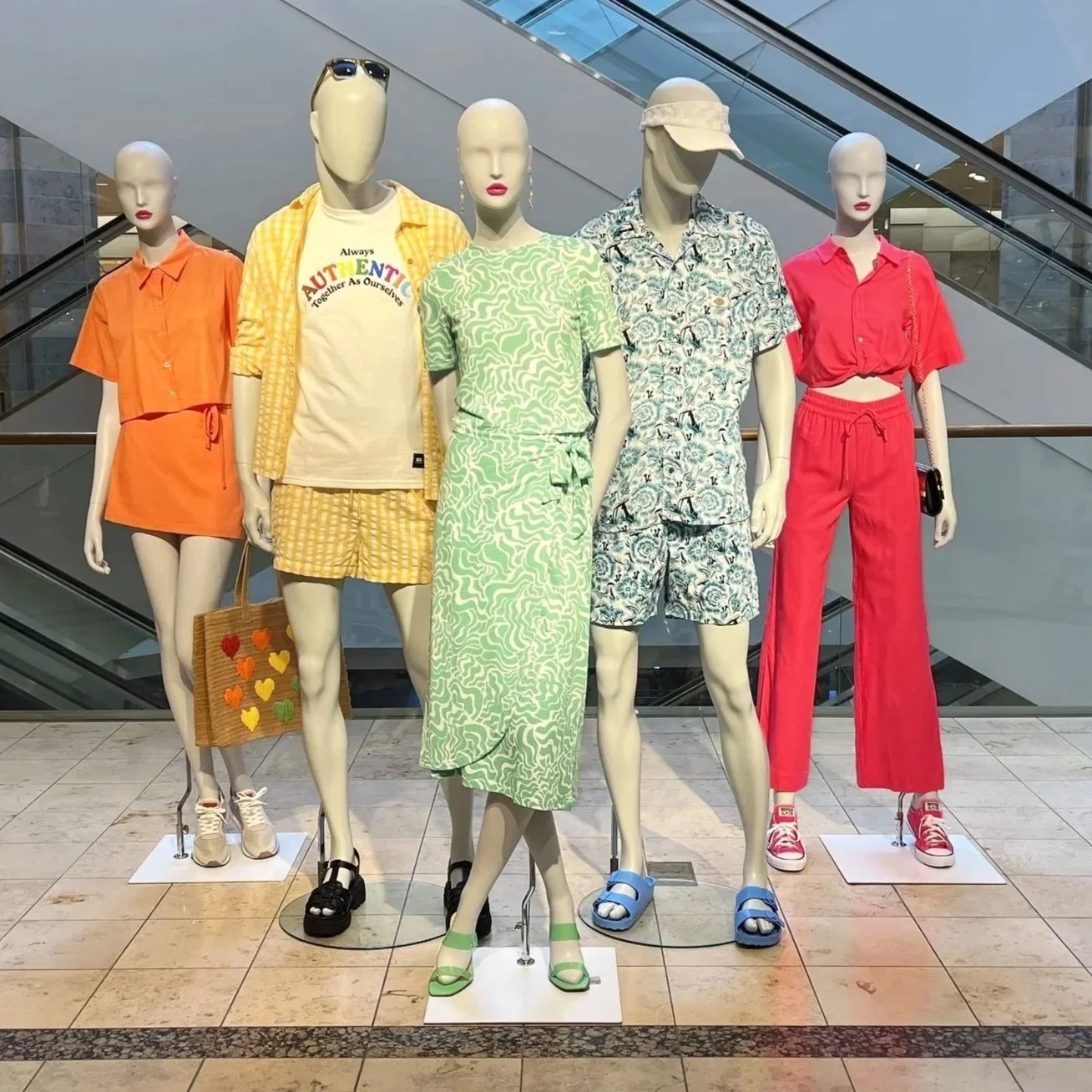 Display of six mannequins dressed in summer casual outfits in a shopping mall. From left to right: orange short-sleeve shirt with matching shorts, sunglasses, and sneakers; yellow checkered shirt with matching shorts, and a straw tote bag with heart designs; light green patterned dress with matching sandals; floral print short-sleeve shirt with matching shorts and blue sandals; bright pink tie-front shirt with matching wide-leg pants and pink sneakers.