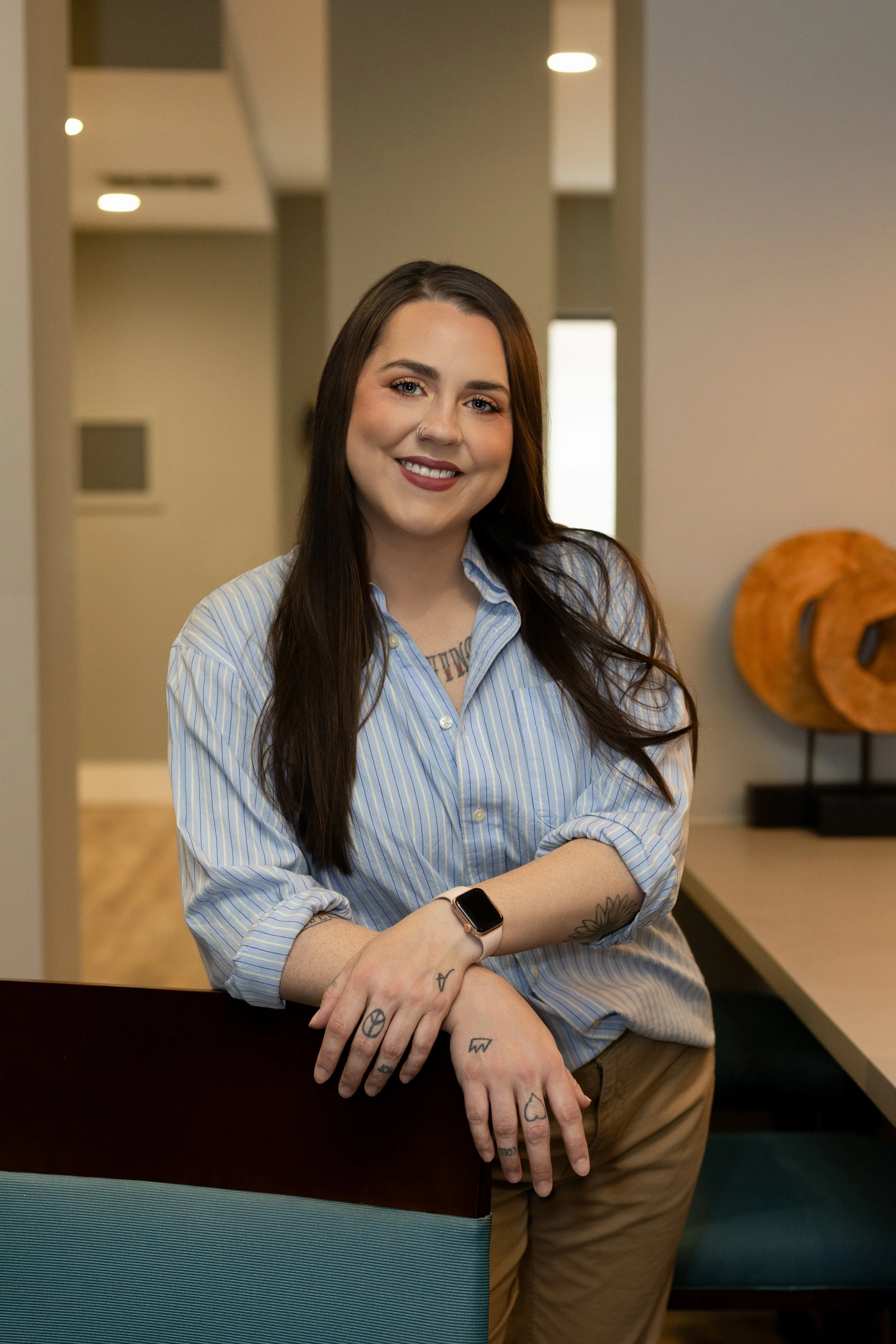 A woman with long dark hair, wearing a blue striped shirt and tan pants, standing in a modern interior space with neutral tones, smiling at the camera.