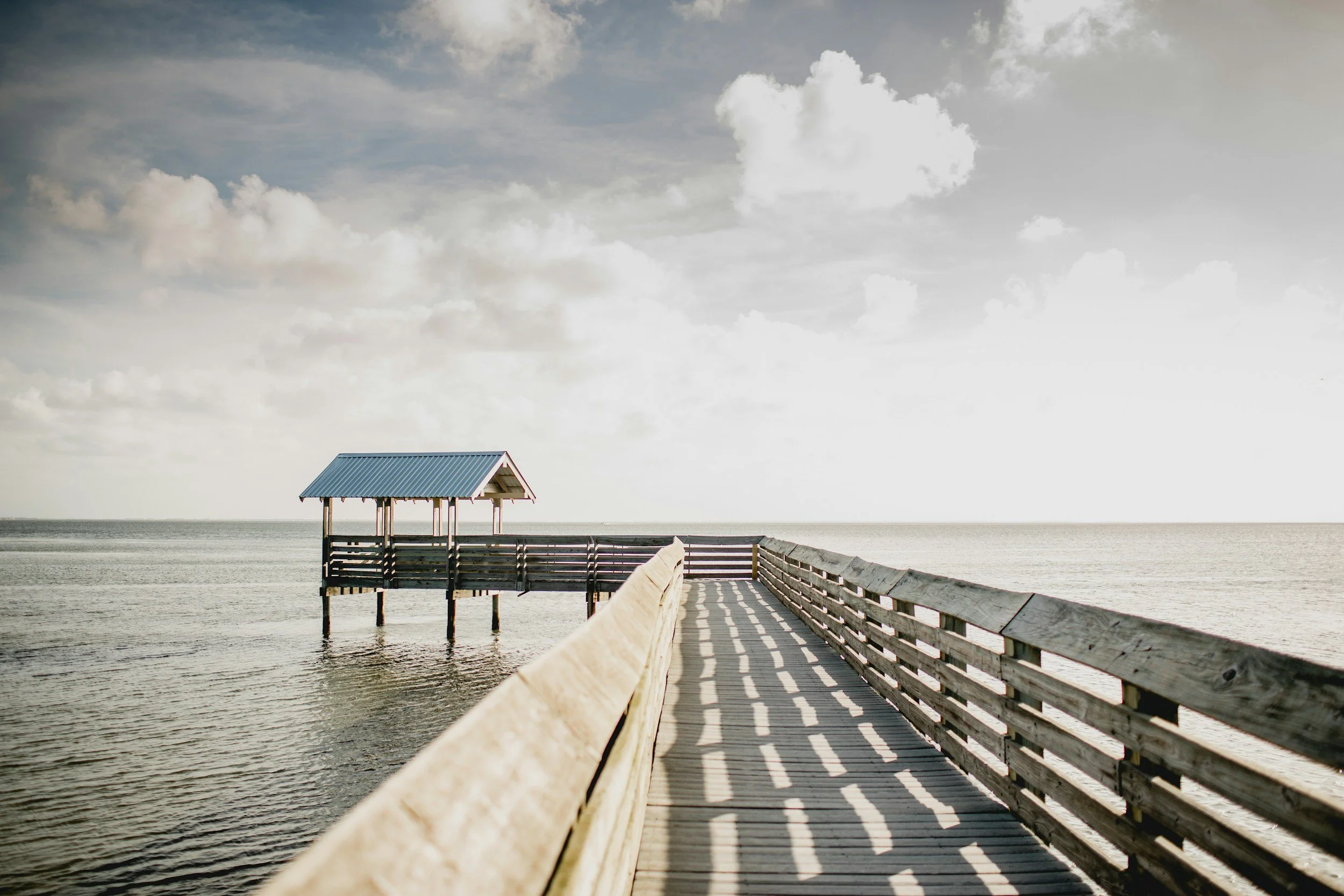 A wooden pier extending into the ocean with a small shelter at the end under a cloudy sky.