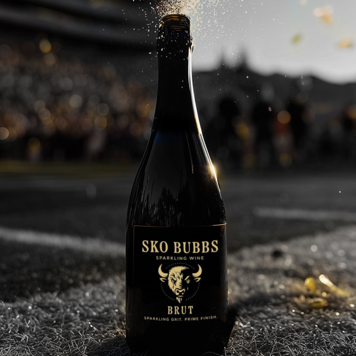 A bottle of SKO Bubbs sparkling wine with a black label, featuring a bull's head graphic, placed on a gravel surface with a blurred outdoor background during sunset.