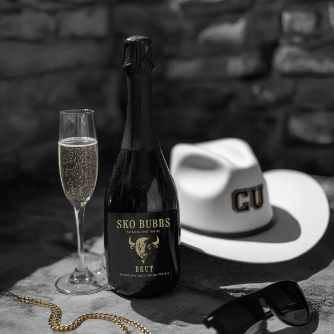 A bottle of SKO BUBBS sparkling wine, a filled champagne flute, a white cowboy hat labeled 'CUB', a pair of black sunglasses, and a gold necklace on a flat surface.