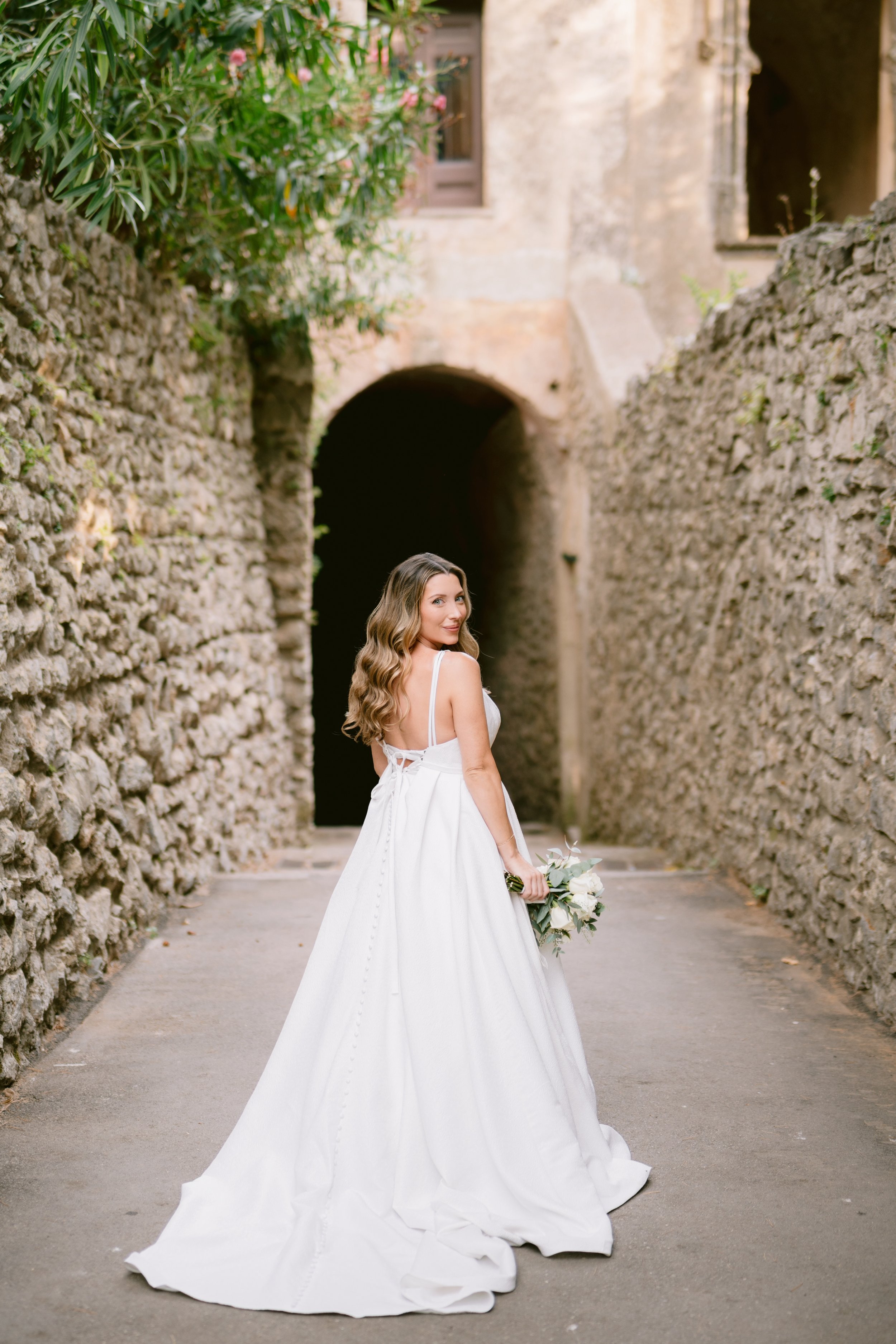 Bride in a white wedding dress holding a bouquet, standing in an outdoor stone alleyway.