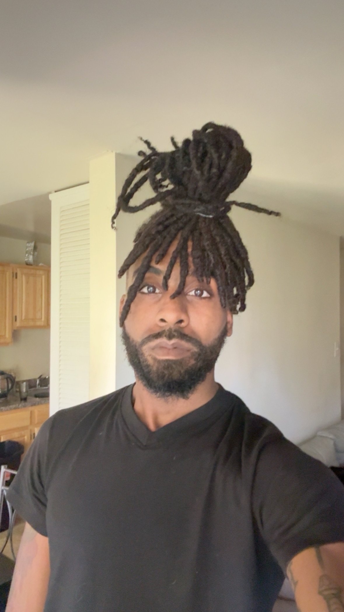 A man with a beard and dreadlocks piled high on his head, wearing a black t-shirt, standing in a room with a kitchen in the background.