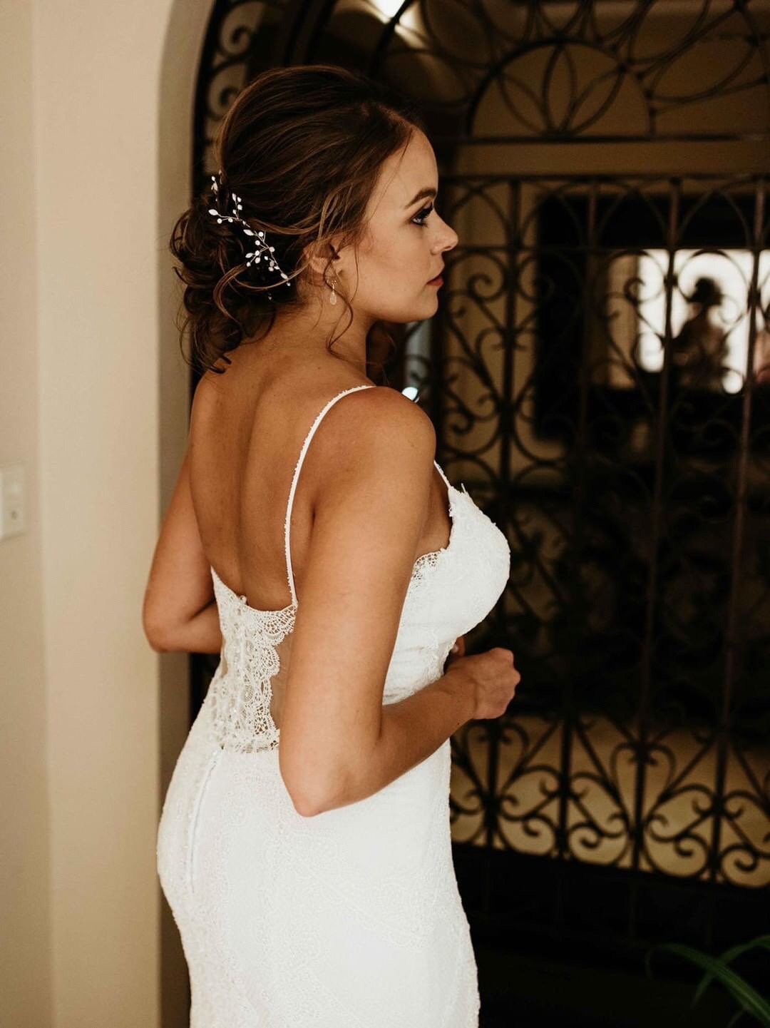 A woman in a white lace wedding dress with spaghetti straps, standing indoors near an ornate metal gate, with her profile turned to the side, and styled hair with a decorative hairpiece.