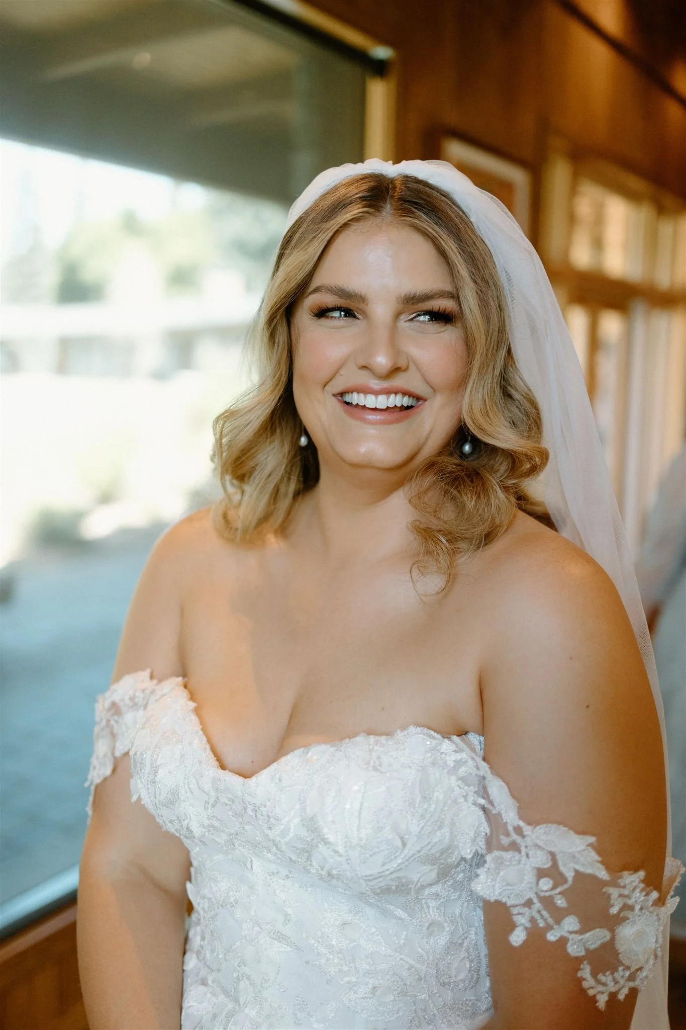 A smiling bride with blonde hair and pearl earrings, wearing a strapless lace wedding gown and veil, posing indoors with a wooden interior and window behind her.