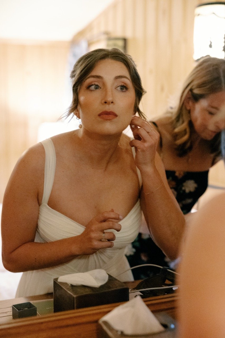 A woman in a white dress with makeup and earrings getting ready, looking into a mirror.