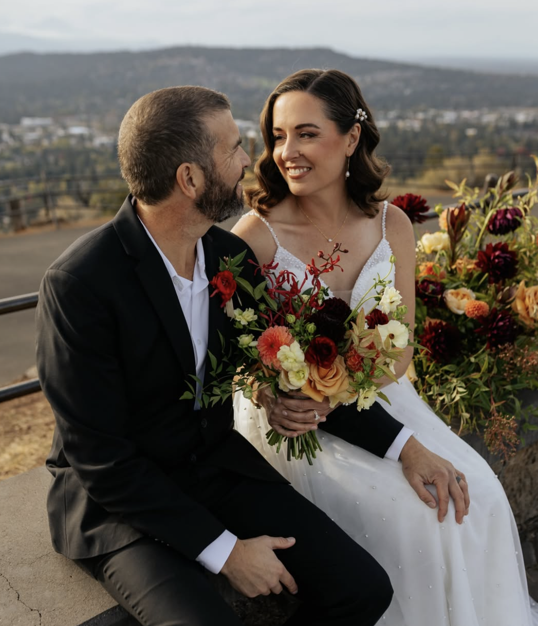 A bride and groom sitting outdoors surrounded by flowers, smiling at each other during their wedding.
