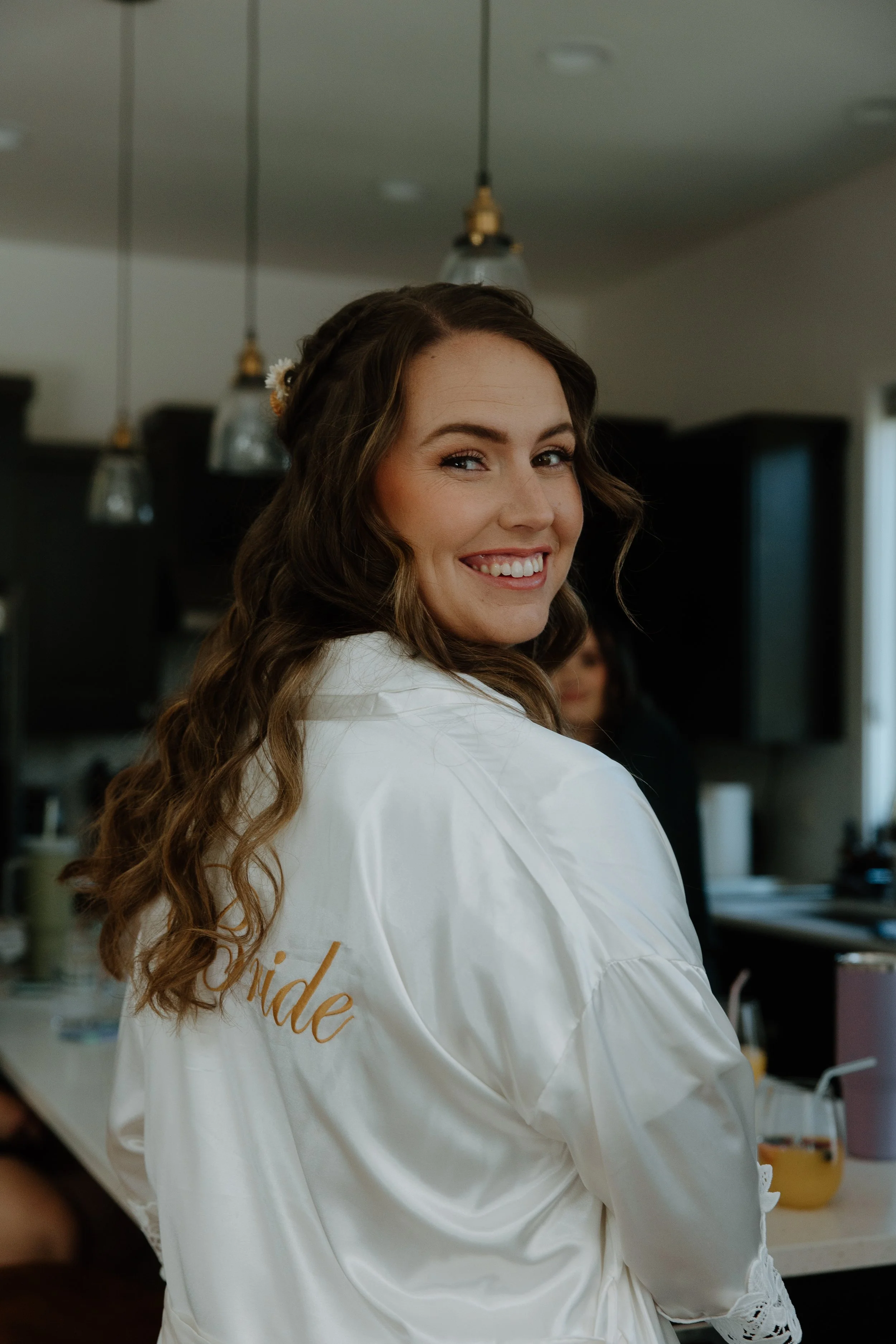 A smiling bride with long, wavy brown hair, wearing a white satin robe with 'bride' written on the back, in a kitchen setting.