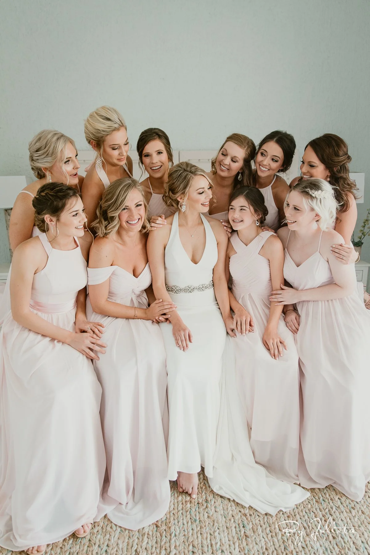 A bride and her eleven bridesmaids sitting together, all dressed in white or blush-colored dresses, smiling and interacting with each other in a well-lit room.