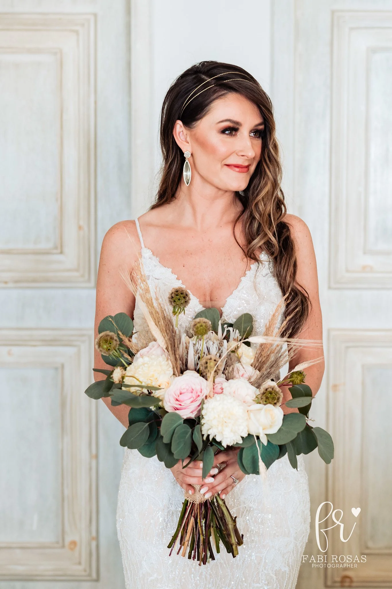 A woman in a white lace dress holding a bouquet of white and pink flowers with greenery and dried grasses, standing in front of paneled white walls.
