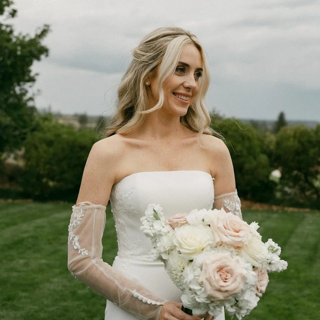 A bride with long blonde hair in a strapless wedding dress holding a bouquet of white and blush pink roses, standing outdoors on a cloudy day.