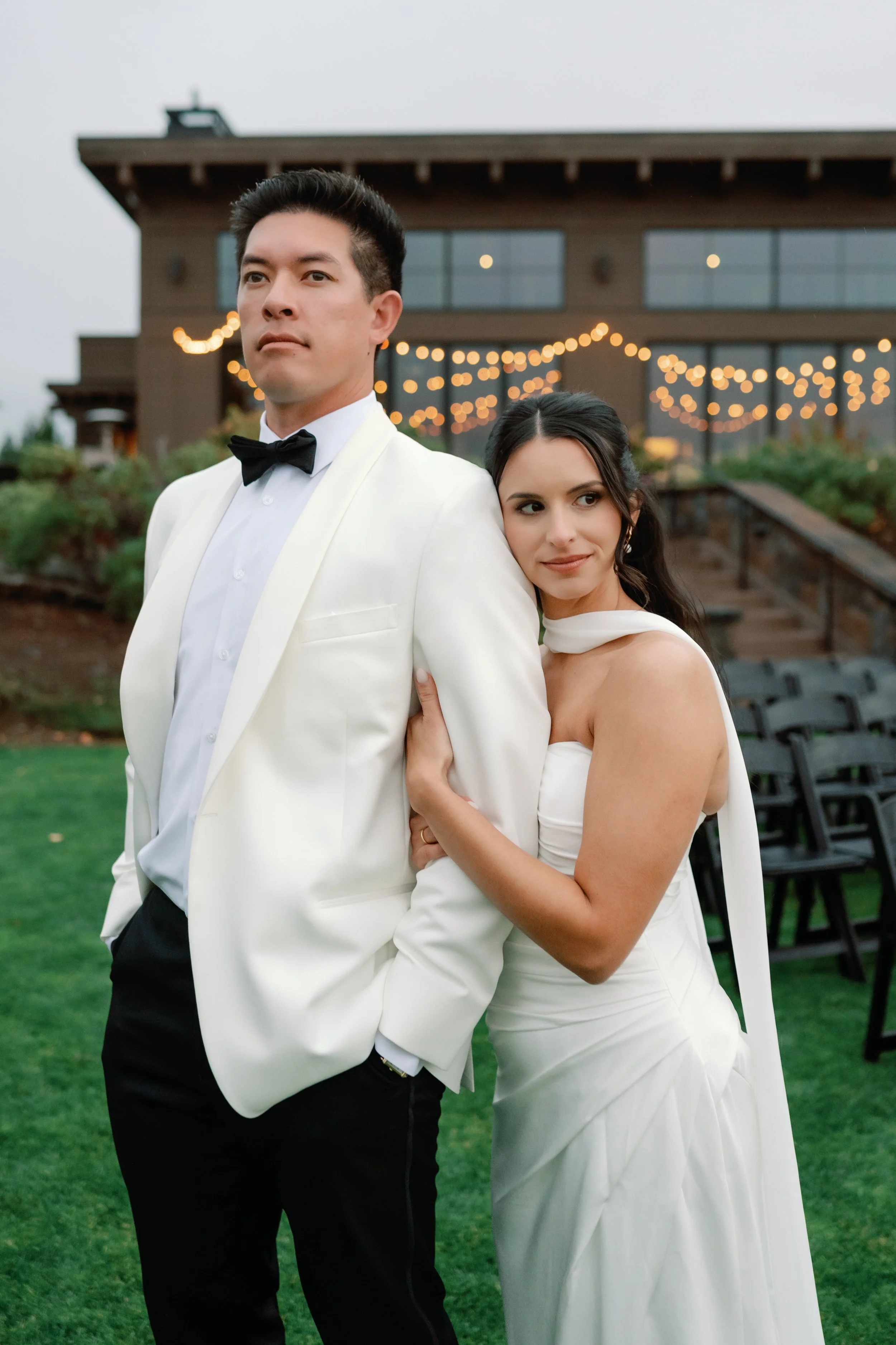 A bride and groom standing outdoors at sunset, with the bride hugging the groom's arm, both dressed in formal wedding attire.