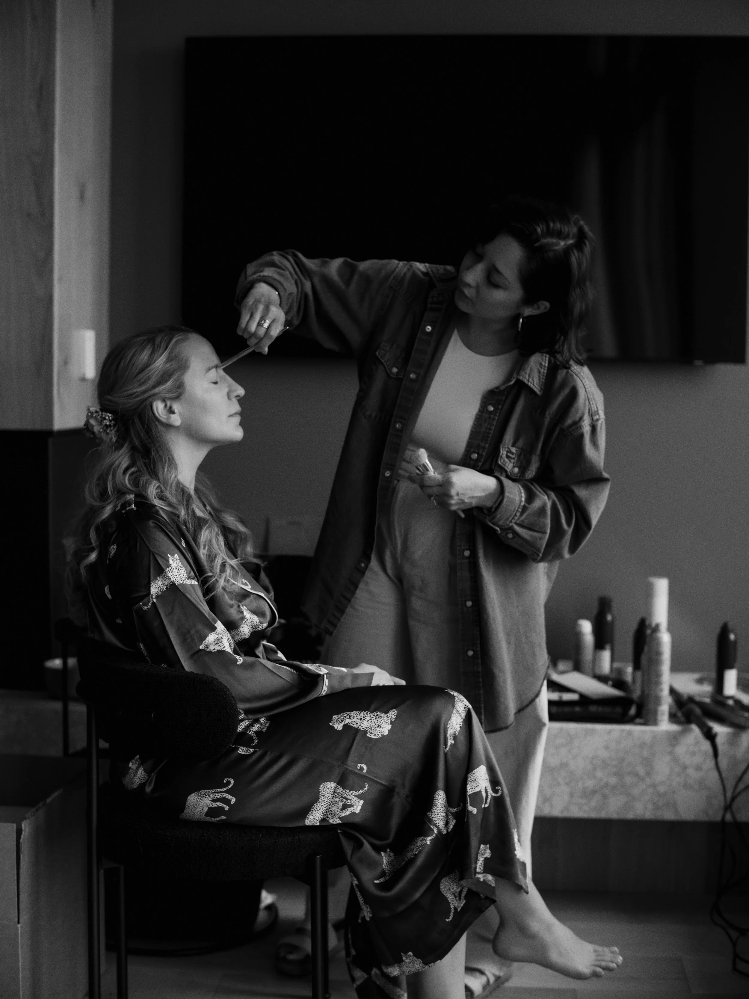 A woman getting her makeup done by another woman in a room with cosmetics on the table.