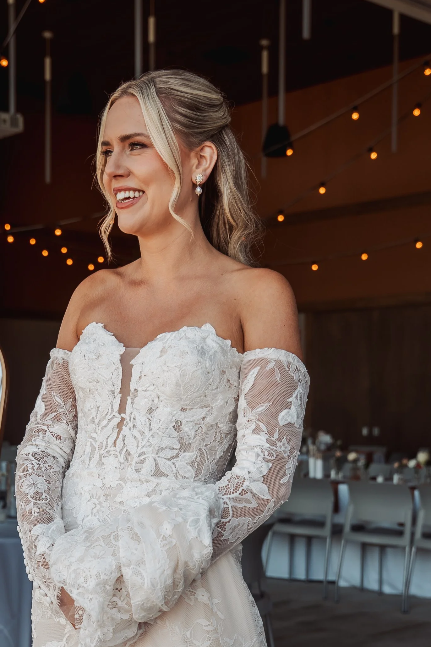 Bride in a lace wedding dress with off-shoulder sleeves, smiling at a wedding reception with string lights overhead and decorated tables in the background.