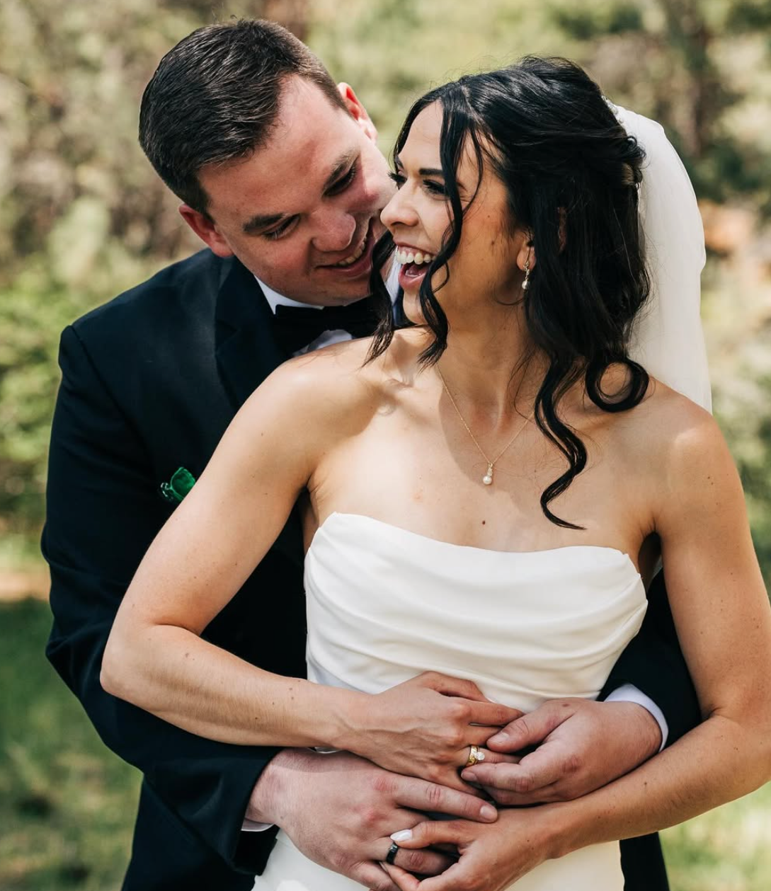 A happy bride and groom share a joyful moment outdoors on their wedding day, embracing each other with the groom wrapping his arms around the bride's waist as they smile and laugh.