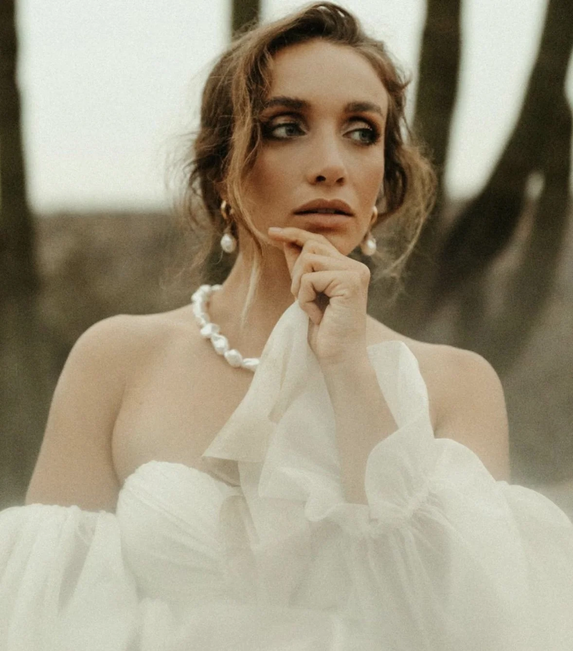 A woman with wavy brown hair, wearing pearl jewelry, holding a white dress or fabric, standing outdoors near trees.