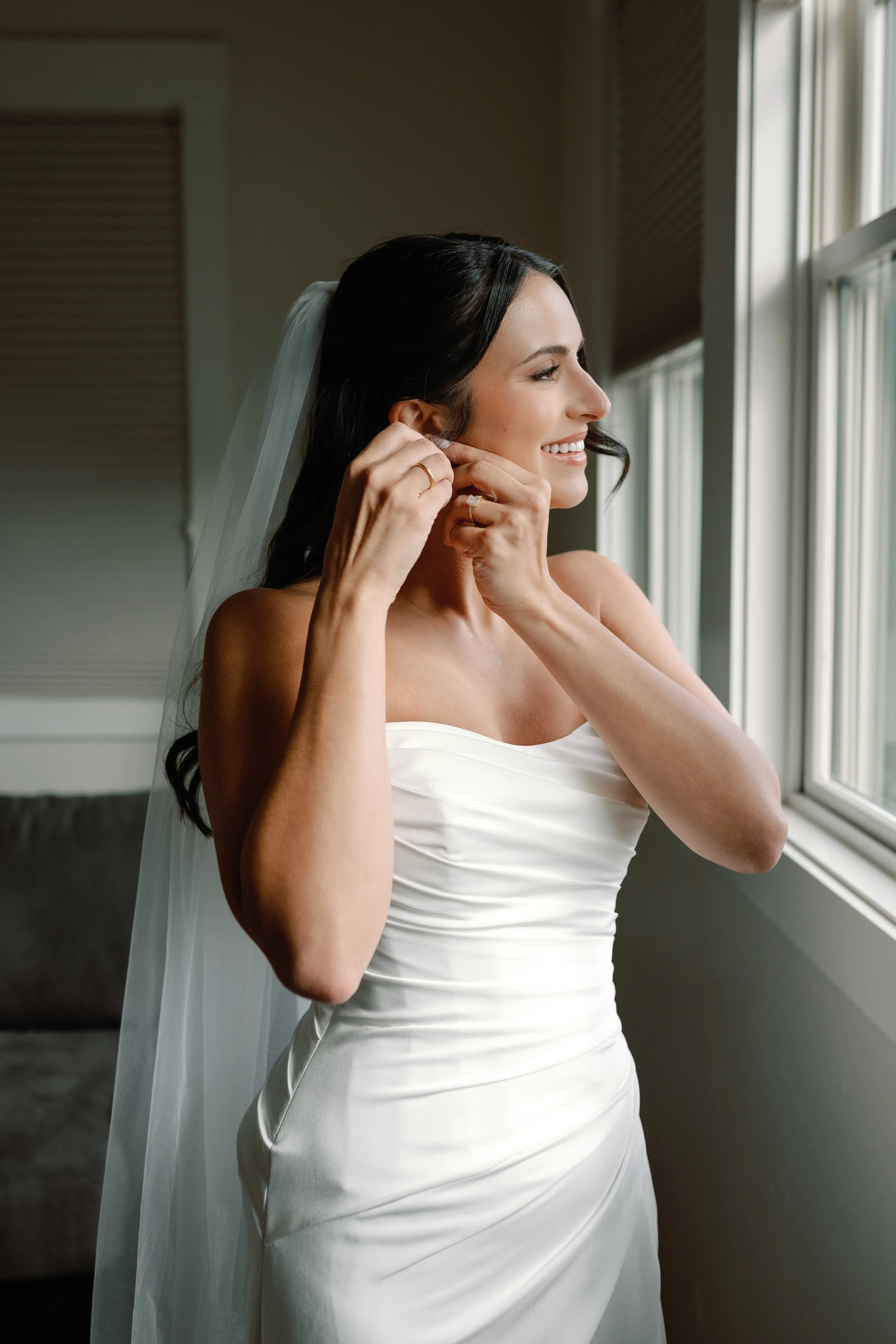 Brunette bride wearing a white wedding dress, putting on earrings by a window, smiling.