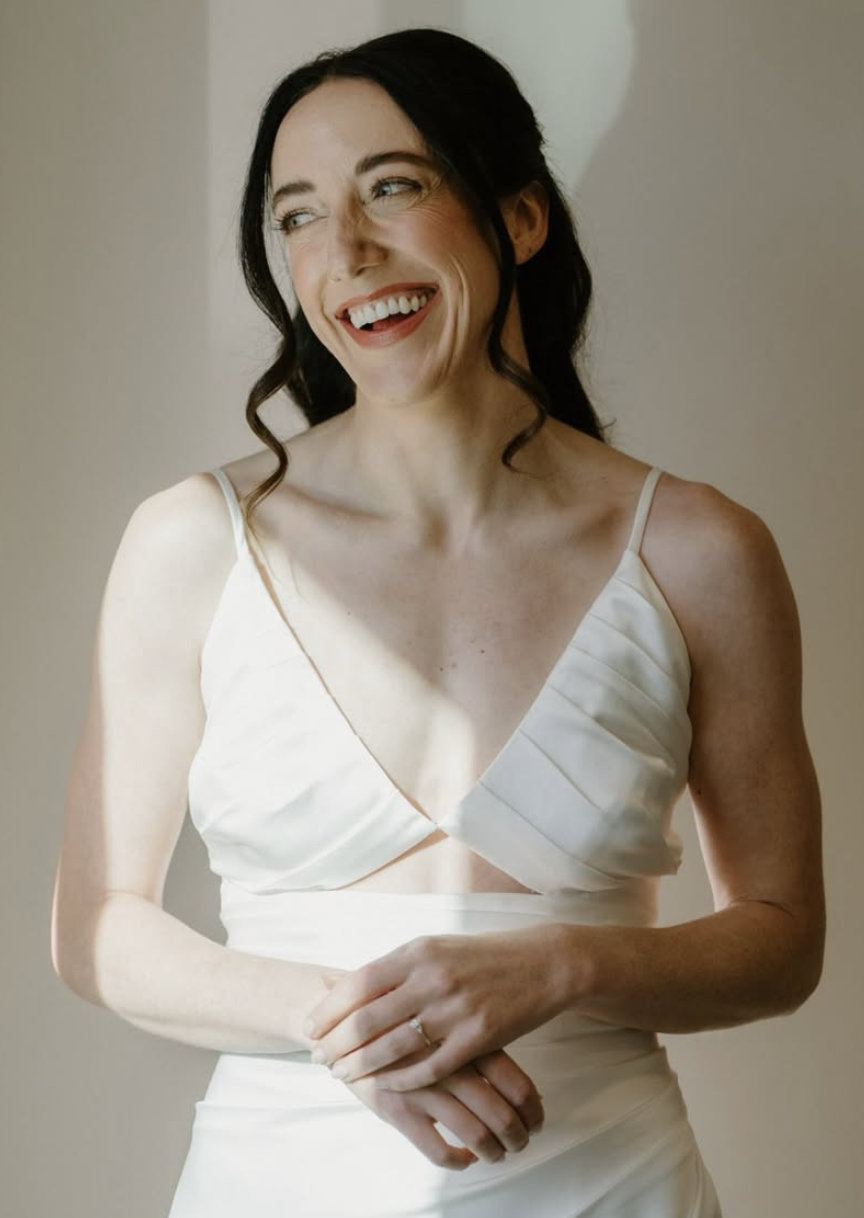 A woman with dark hair styled in loose waves wearing a white dress with thin straps, smiling and looking to her right.
