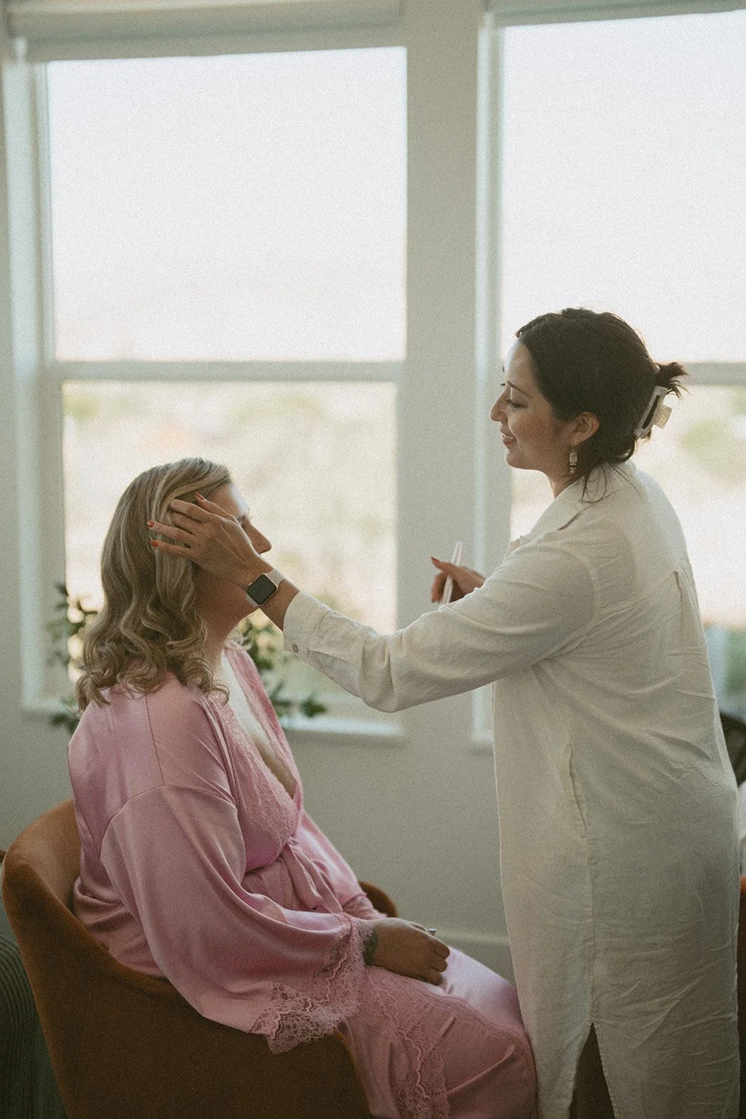 A woman in a pink robe sits in a brown chair while another woman in a white coat applies makeup or skincare to her face in front of a large window with sunlight streaming in.
