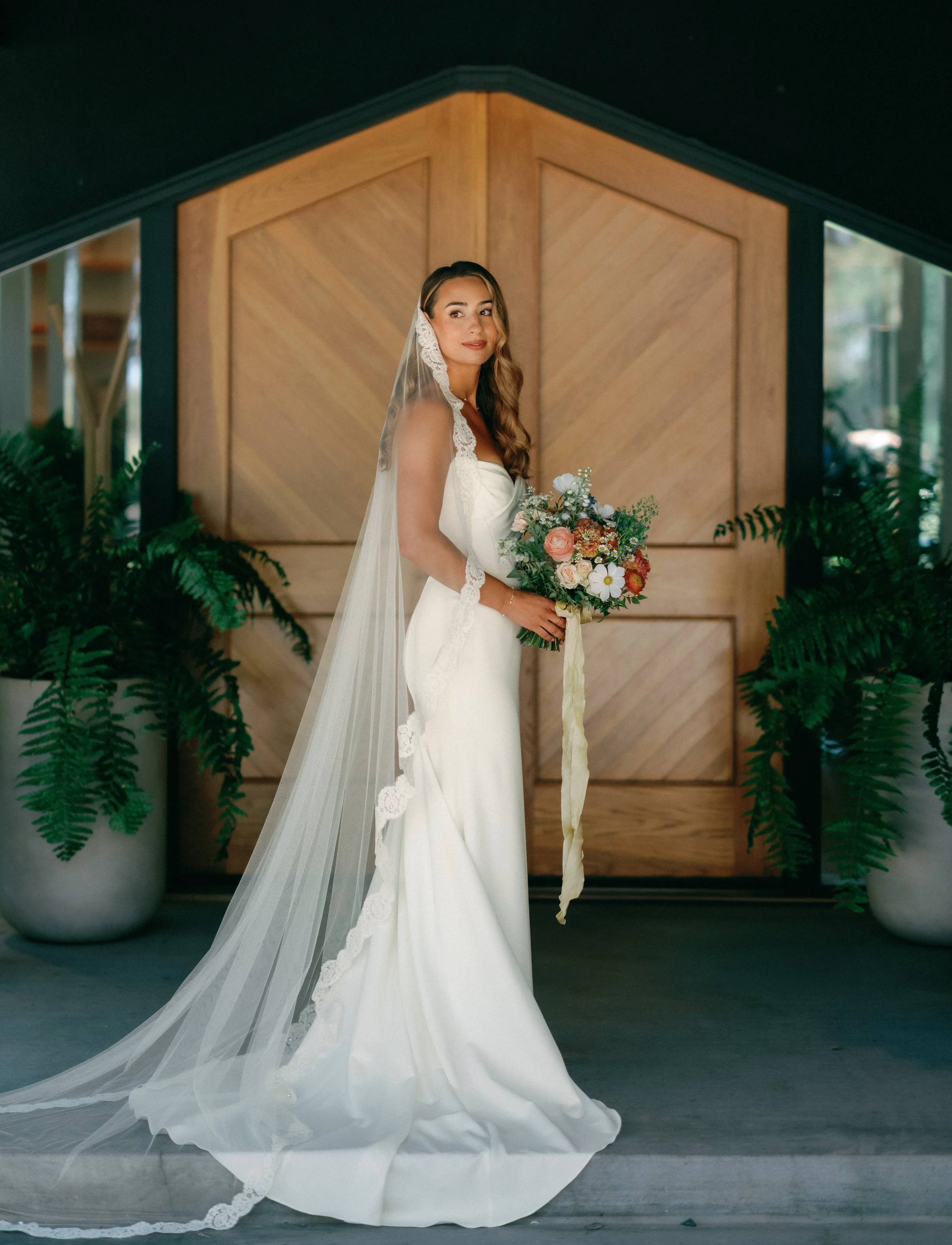 Bride in a white wedding gown with a long veil holding a colorful bouquet standing in front of wooden double doors flanked by large potted ferns.