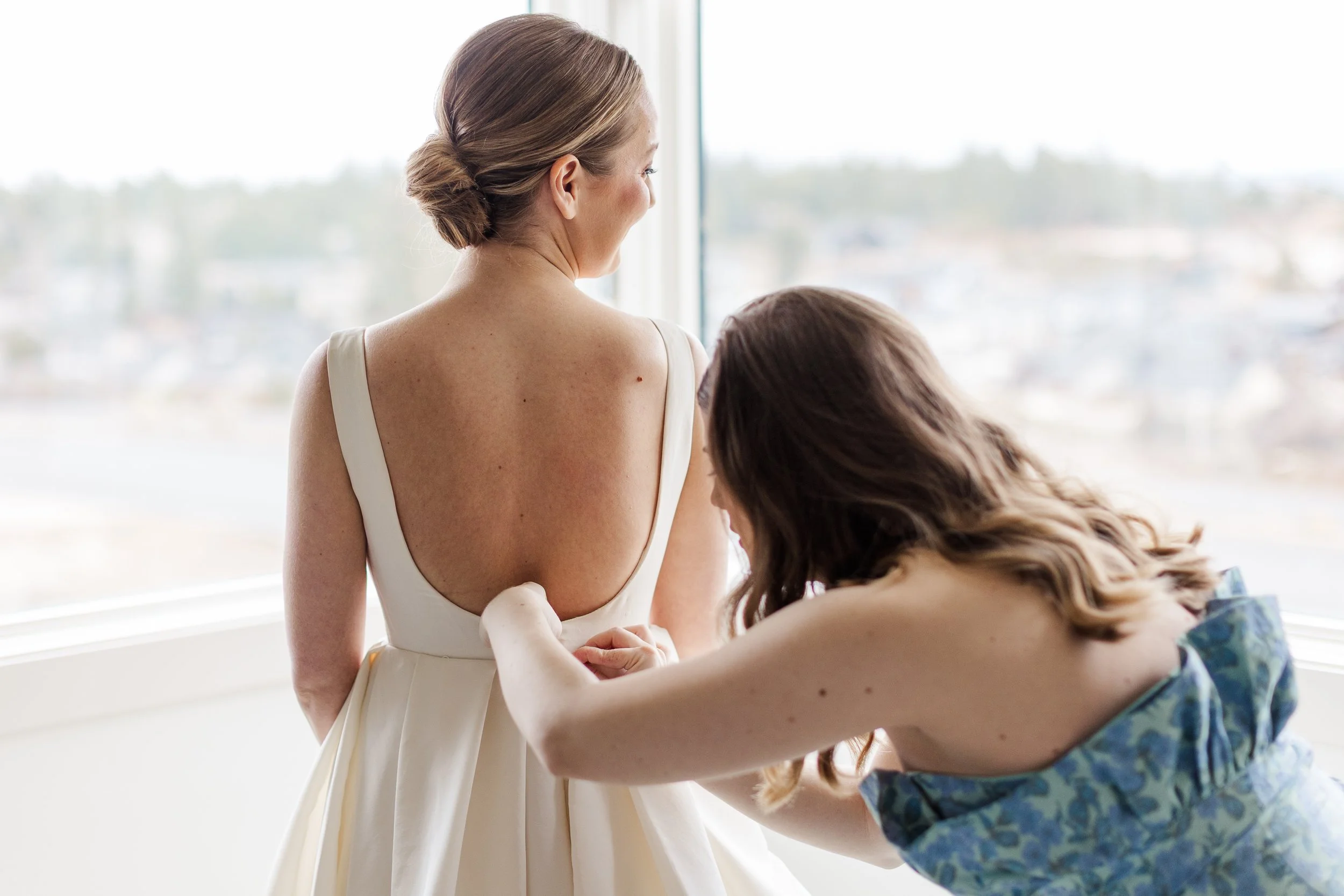 A woman in a white dress having her sleeve adjusted by another woman in a blue floral dress, near a large window with a city view.