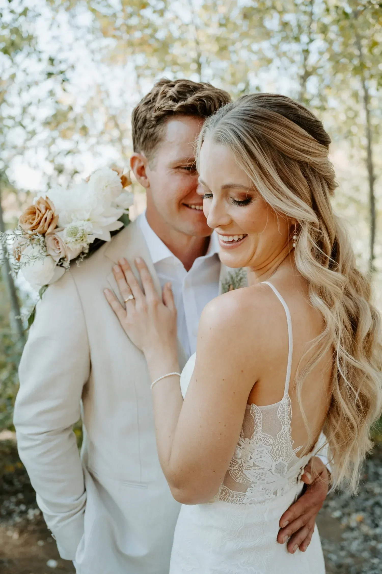 A bride and groom smiling close together outdoors during their wedding, with trees and foliage in the background.