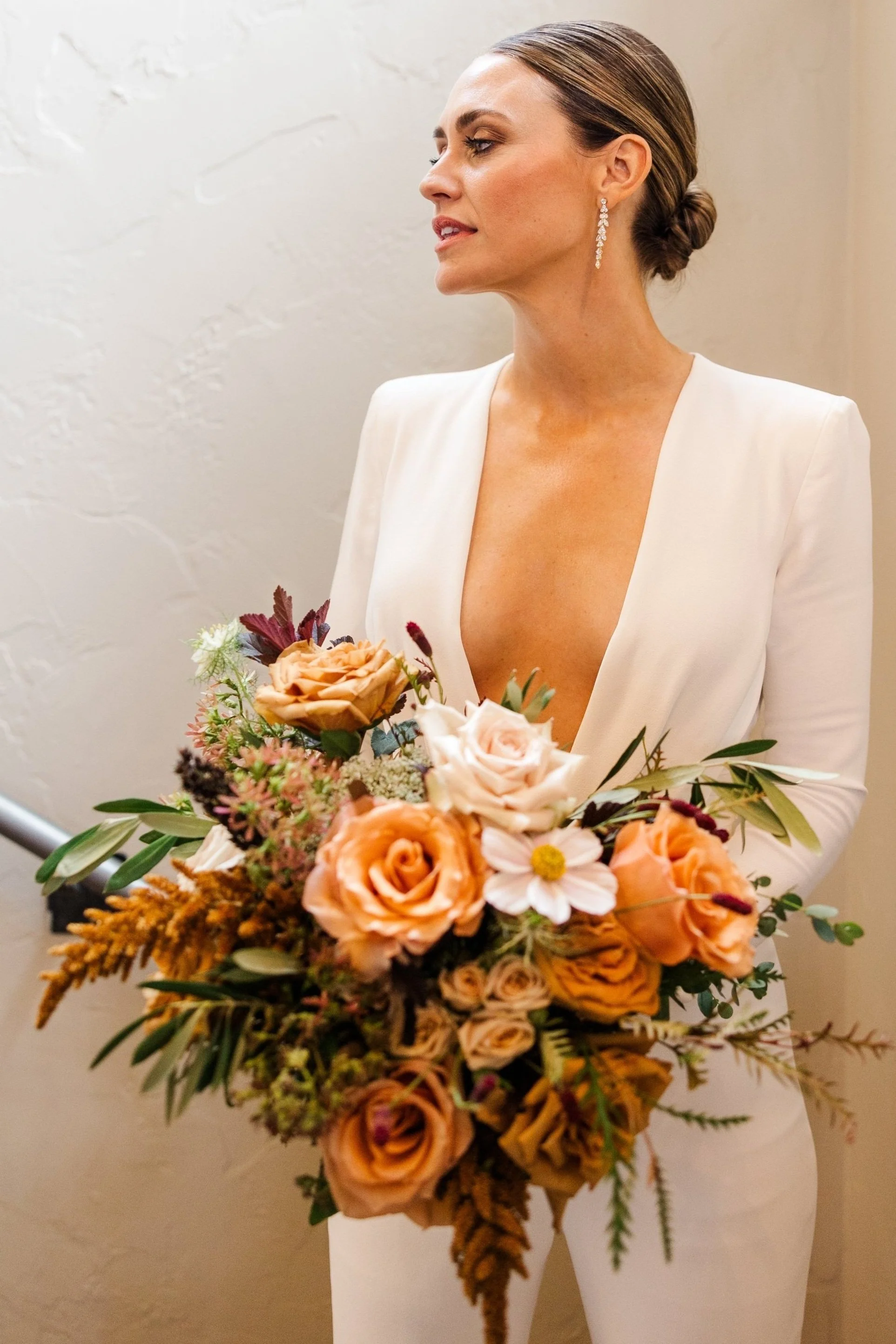 Woman in a white blazer holding a bouquet of peach, pink, and white flowers, with her hair styled in an elegant bun and wearing long earrings.