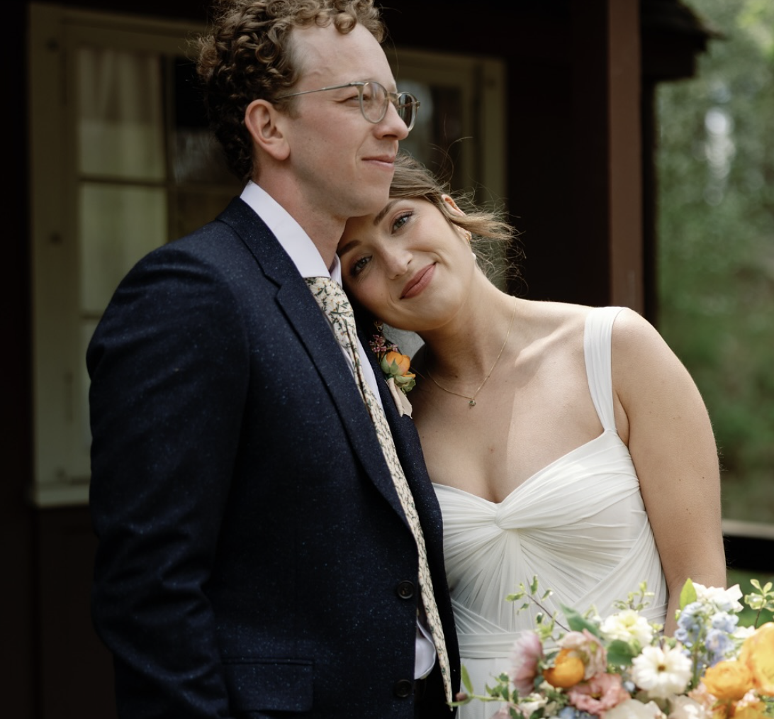 A happy bride and groom on their wedding day, sharing a tender moment outdoors. The groom is wearing glasses and a dark suit, while the bride leans her head on his shoulder, smiling softly with a bouquet of flowers in front of her.
