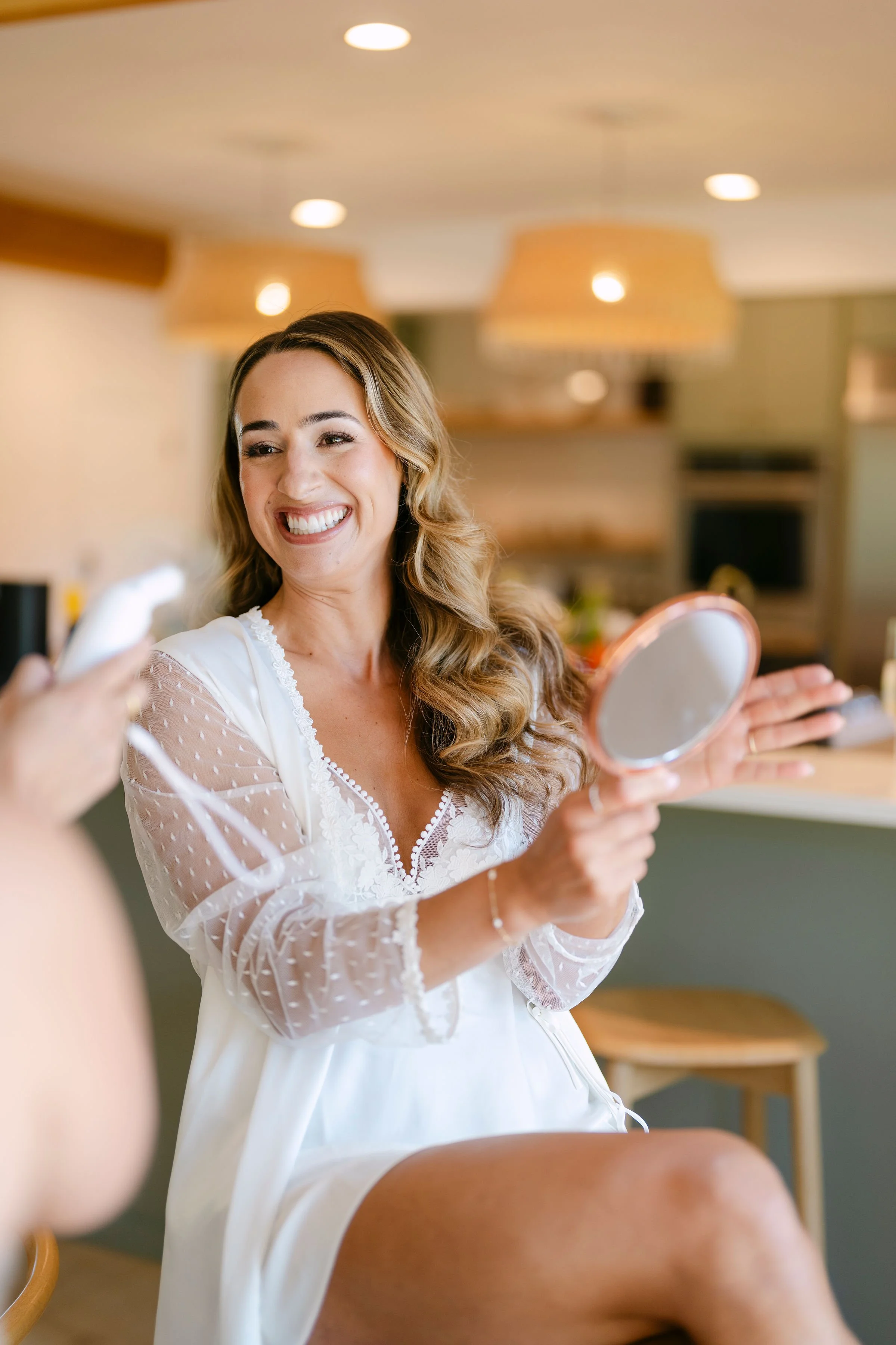 A woman smiling and looking into a handheld mirror in a brightly lit room, possibly preparing for an event.