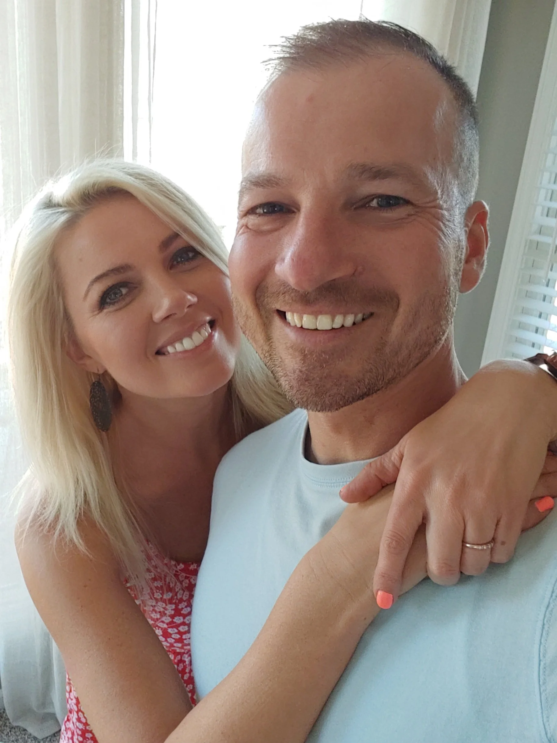 A smiling couple taking a selfie together indoors with sunlight coming through a window behind them.