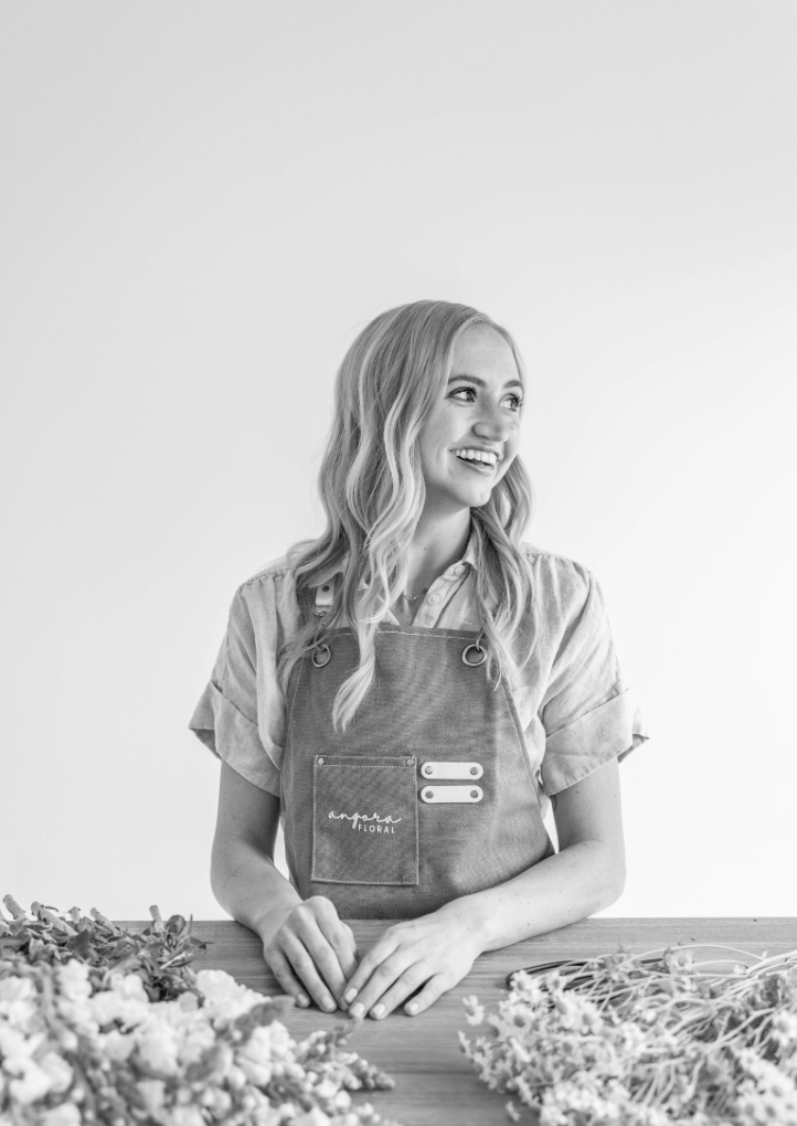A young woman in an apron working as a florist at a table with flowers, smiling and looking to the right in a bright, plain background.