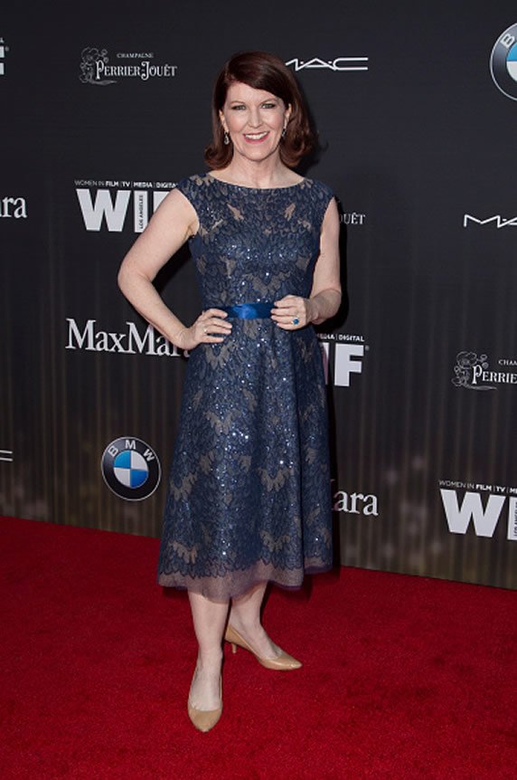 A woman in a navy blue lace dress standing on a red carpet at an event. She has shoulder-length red hair and is smiling at the camera.