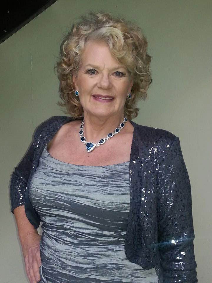 Older woman with curly blonde hair wearing a sparkly black jacket, a patterned gray top, and blue jewelry, smiling against a light green wall.