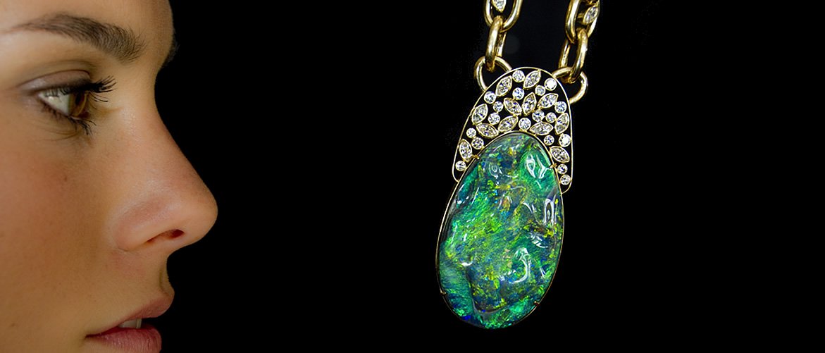 Close-up of a woman looking at a large, oval-shaped opal and diamond pendant necklace against a black background.