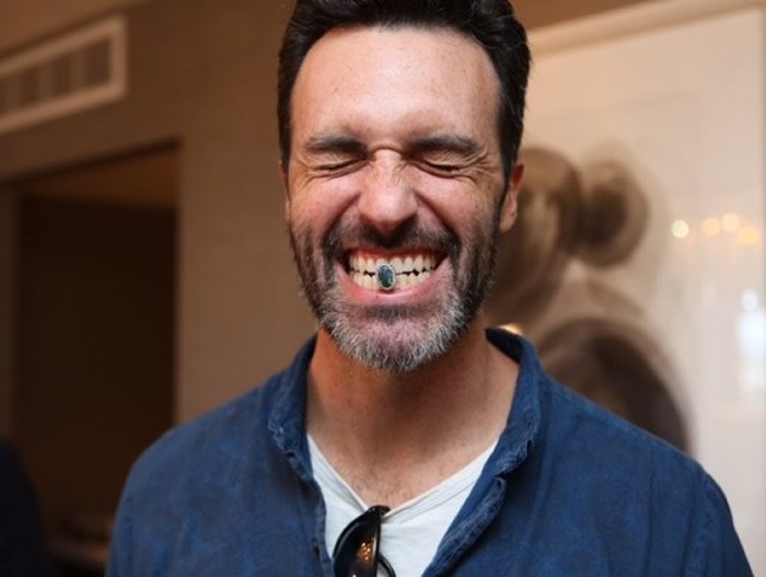 Man with dark hair and beard smiling with eyes closed, showing a tooth with a gold grill and a turquoise stone, wearing a blue button-up shirt, in an indoor setting.