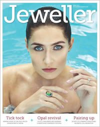 Close-up of a woman with blue eyes and dark hair styled back, holding a ring with a large green gemstone, on the cover of Jeweller magazine.