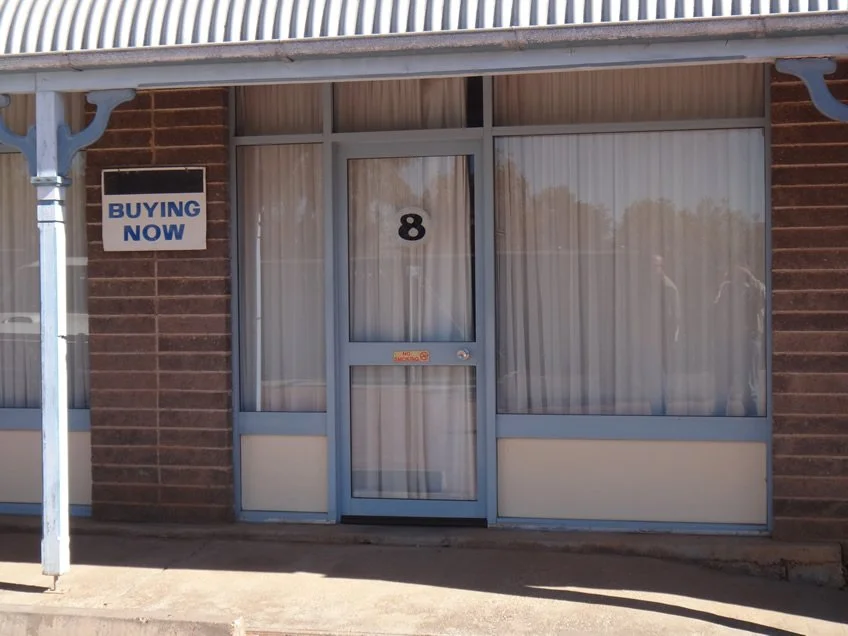 Storefront with a glass door and windows framed in blue, brick walls, a sign that reads 'BUYING NOW' in blue and white, and the number 8 on the door.