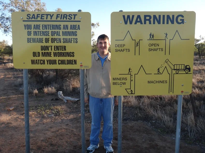 Man standing between two yellow warning signs at an opal mining site, surrounded by dry grass and trees.