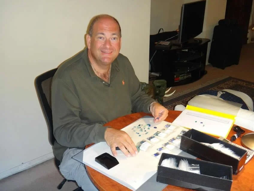 A man sitting at a table with various small items, such as gemstones, tools, and paperwork, in a room with a television and TV stand in the background.