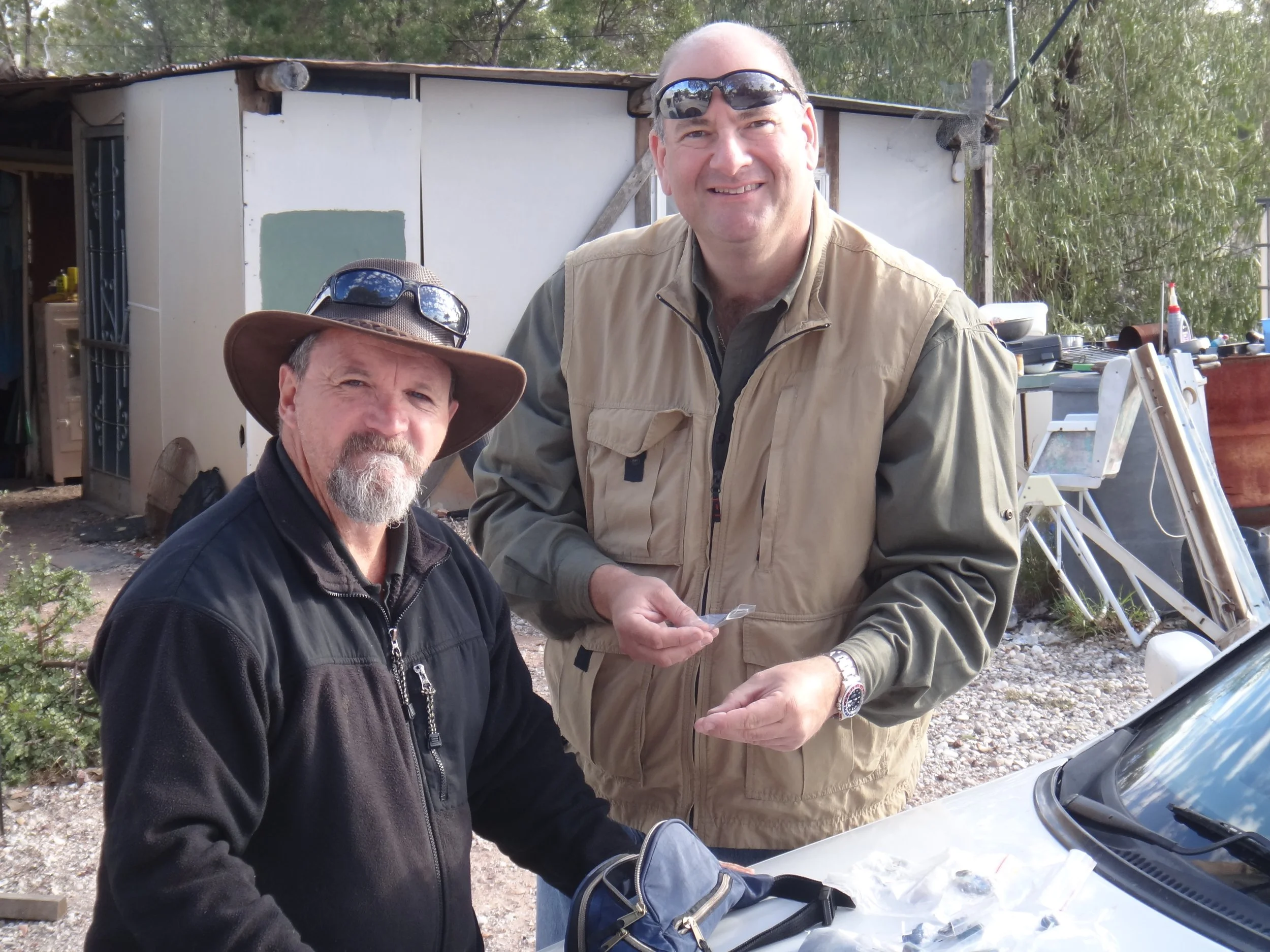 Two men, one with a hat and beard, the other on the right wearing glasses on his head, are standing outdoors near a car with tools and equipment in the background.