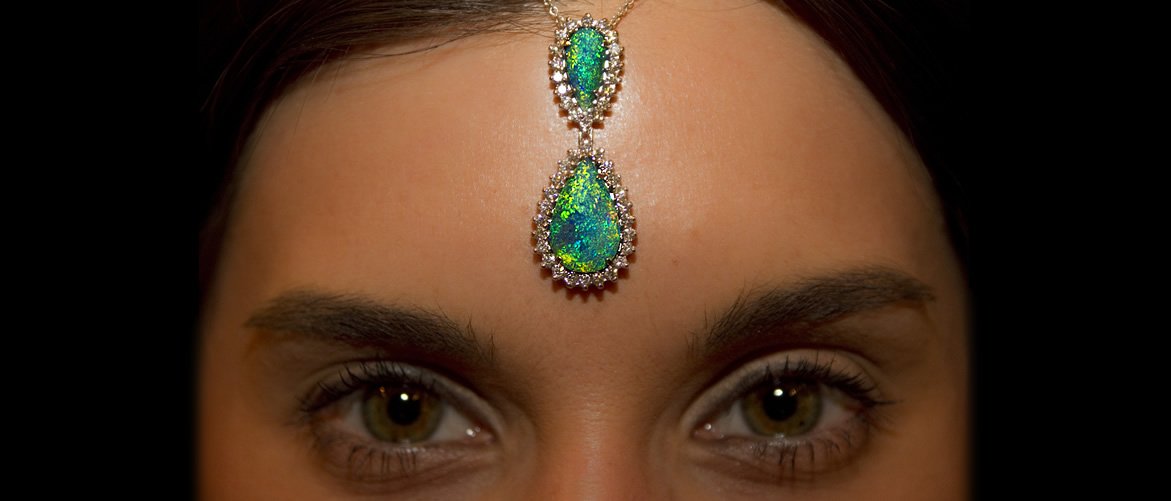 Close-up of a woman's face focusing on her eyes, with a decorative jeweled headpiece featuring green and blue iridescent stones hanging on her forehead.